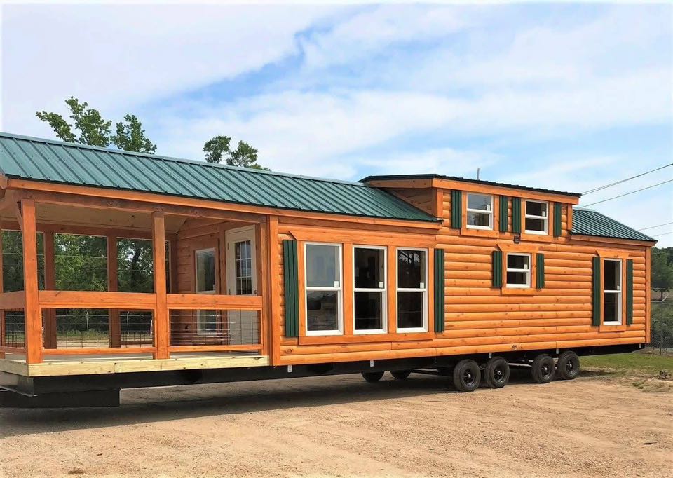 Log cabin-style mobile home with a green metal roof on multiple wheels. It features large windows and a covered porch, set against a blue sky.
