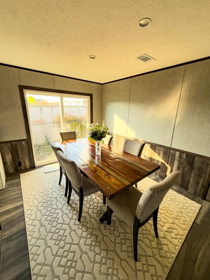 Dining room with a rustic wooden table, six upholstered chairs on a patterned rug. Sunlight streams through a sliding glass door, creating a cozy ambiance.
