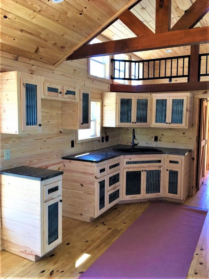 Rustic kitchen with light wood cabinetry and black countertops. Exposed beams and wood walls create a cozy atmosphere. A small loft is visible above.
