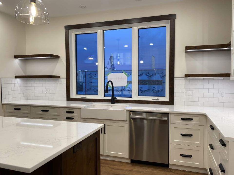 Modern kitchen with white cabinets, sleek black handles, and a stainless steel dishwasher. A large window offers a view of a dusky sky. Warm lighting creates a cozy atmosphere.