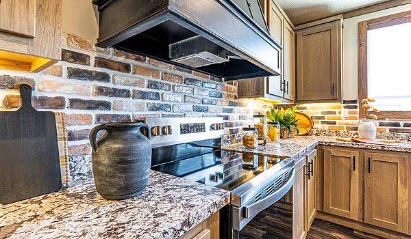Modern kitchen with warm tones, featuring a granite countertop, brick backsplash, and wooden cabinets. Decor includes a black jug and small plants, creating a cozy ambiance.