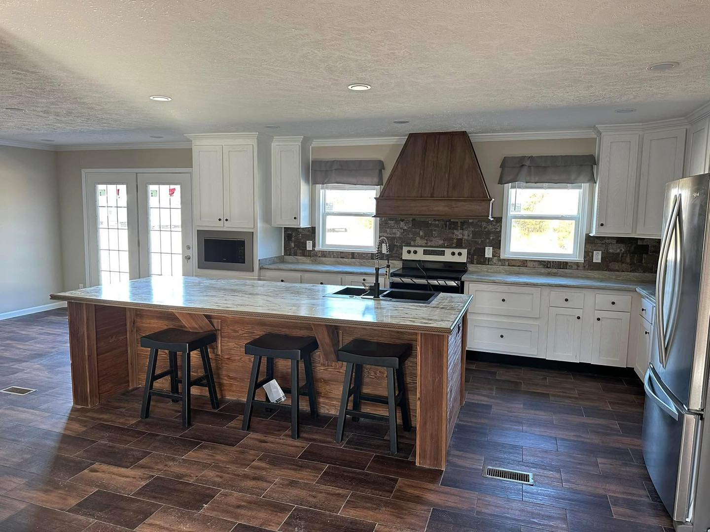 Modern kitchen with white cabinets, dark wood flooring, and a central island with three black stools. Features stainless steel appliances and a warm, inviting tone.
