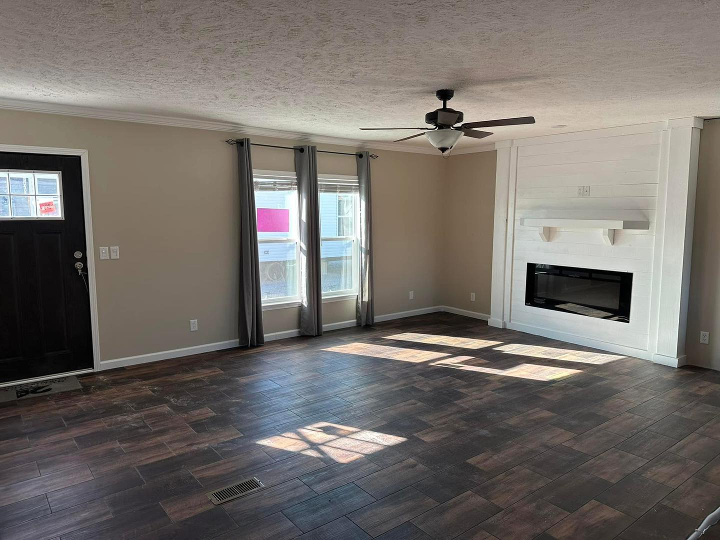 Spacious living room with dark wood floors, beige walls, and a white shiplap fireplace. Two windows with gray curtains and a ceiling fan add elegance.