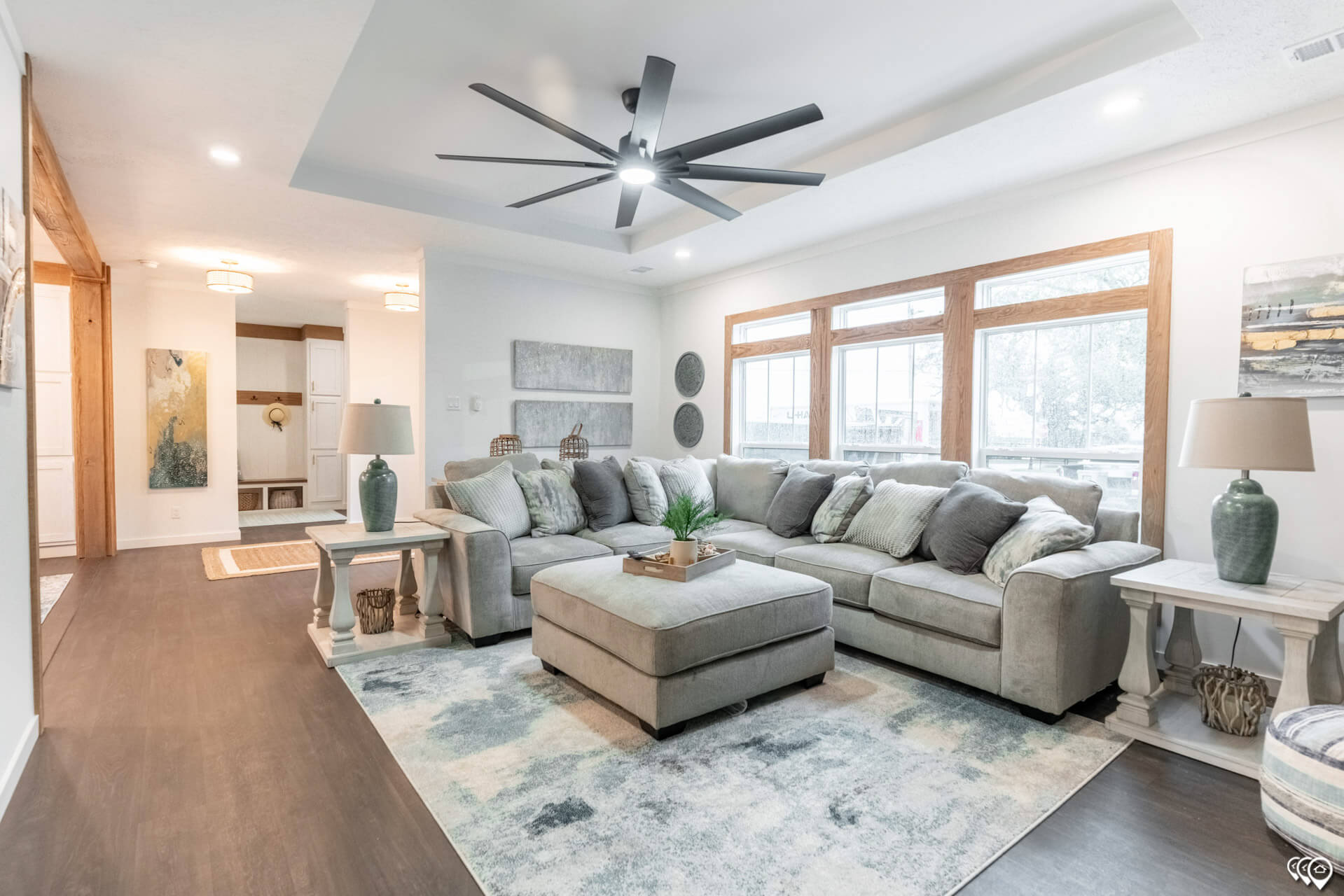 Modern living room with a large gray sectional sofa and ottoman, accented by cozy pillows. Large windows provide natural light, and a sleek ceiling fan adds style. Neutral tones and minimalist decor create an inviting atmosphere.