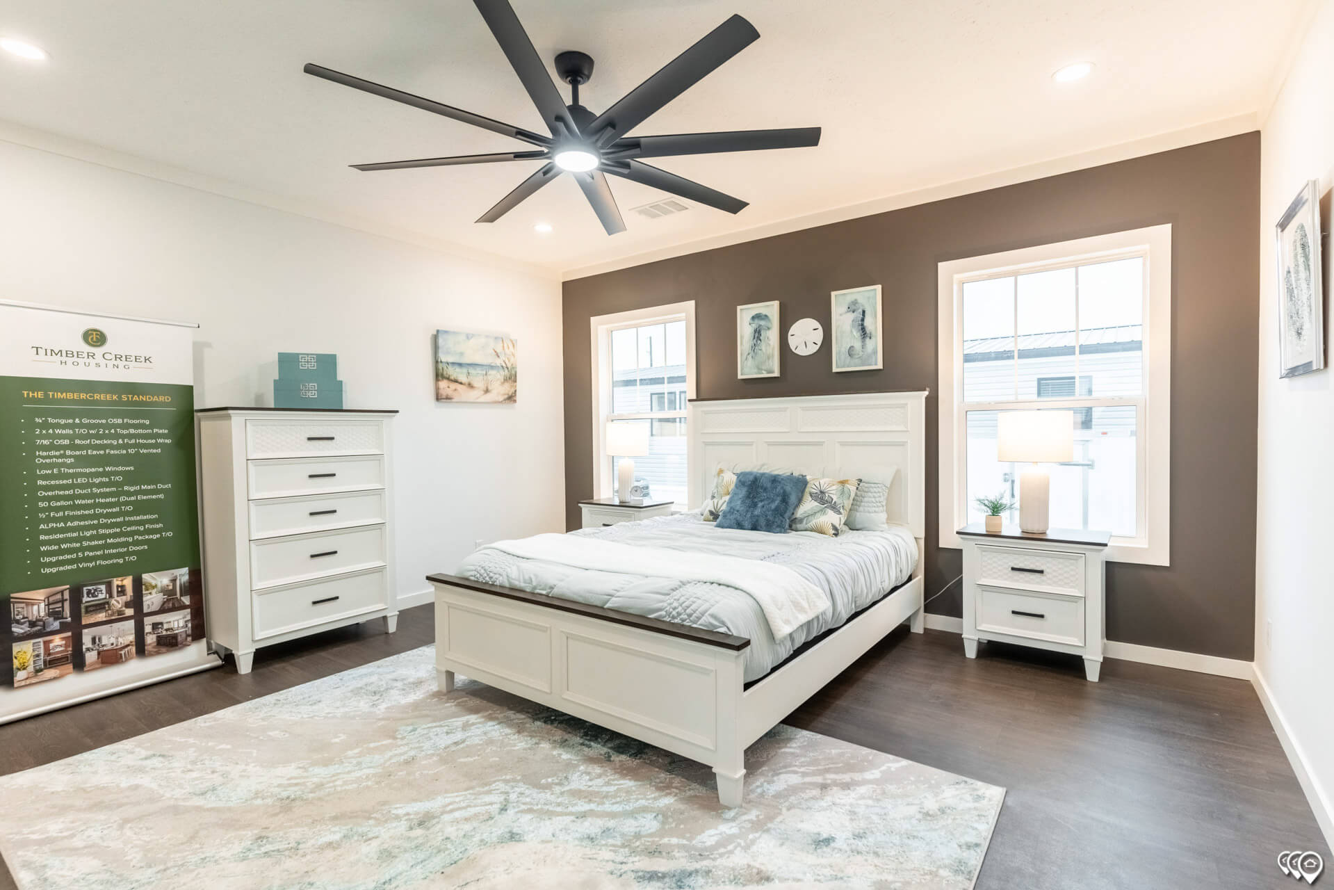 Modern bedroom with a white wooden bed, blue and gray bedding, and matching nightstands. A large dark ceiling fan and warm lighting create a cozy feel.