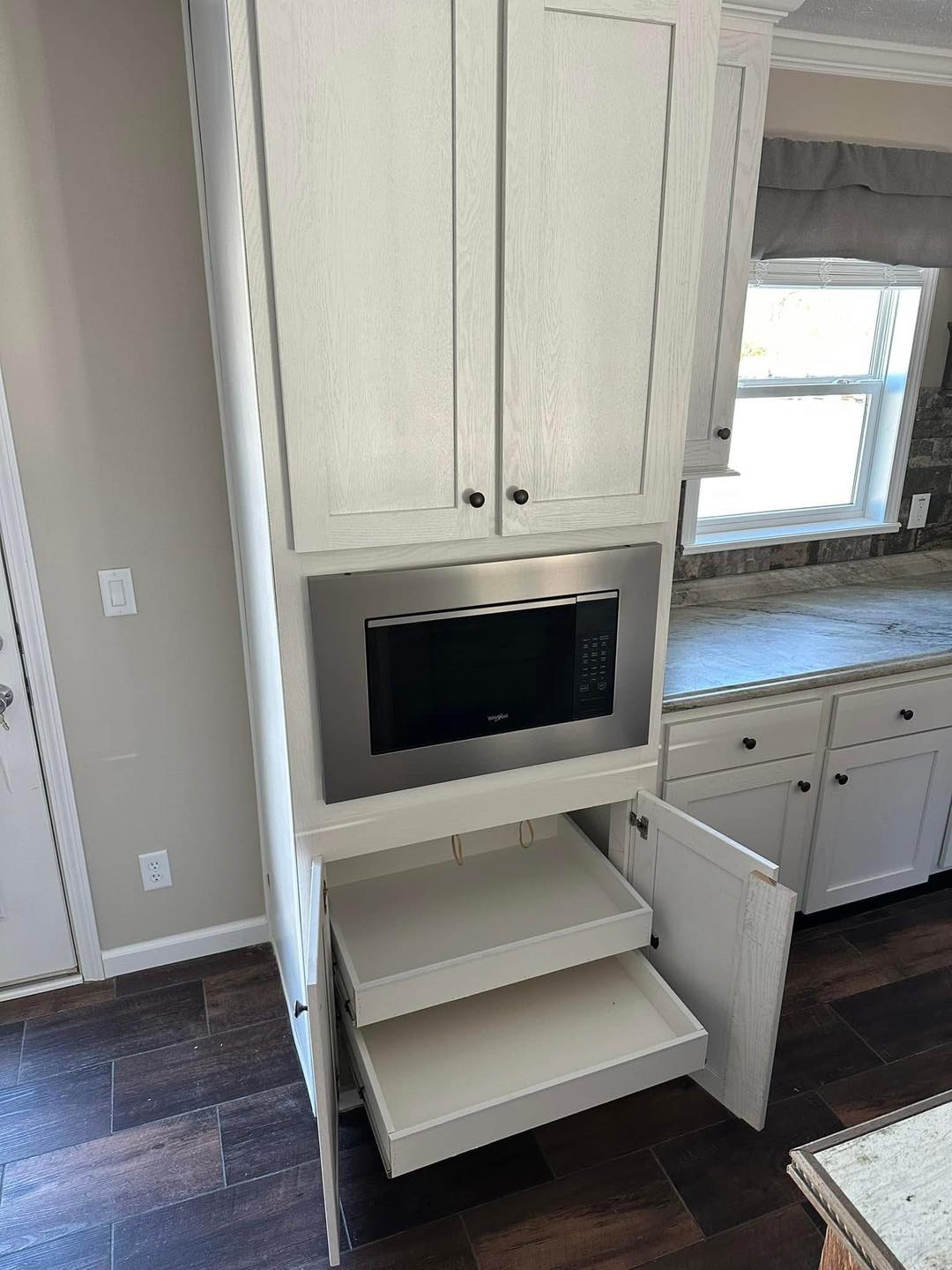 Tall white kitchen cabinet with a built-in microwave and two open pull-out drawers below. A window and countertop are visible in the background. Warm and organized kitchen setting.