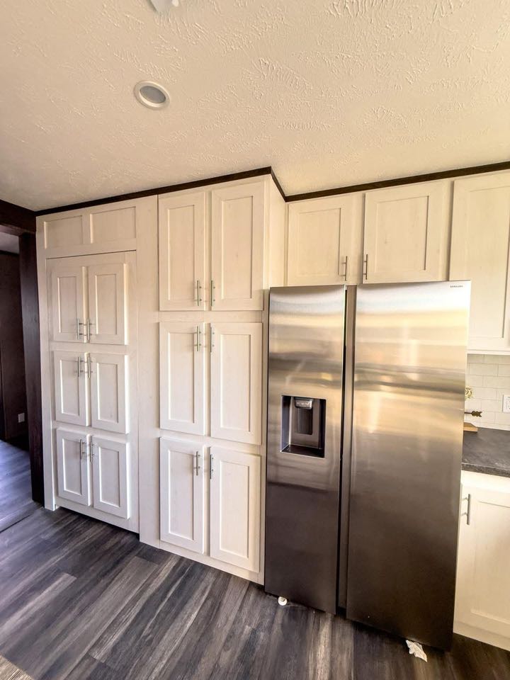 Modern kitchen features sleek stainless steel refrigerator, surrounded by tall white cabinets with silver handles. Dark wood floor adds contrast.