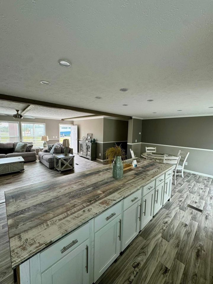 Spacious open-plan living area with rustic wooden flooring and a large kitchen island in the foreground. Cozy sitting area with sofas and natural light.
