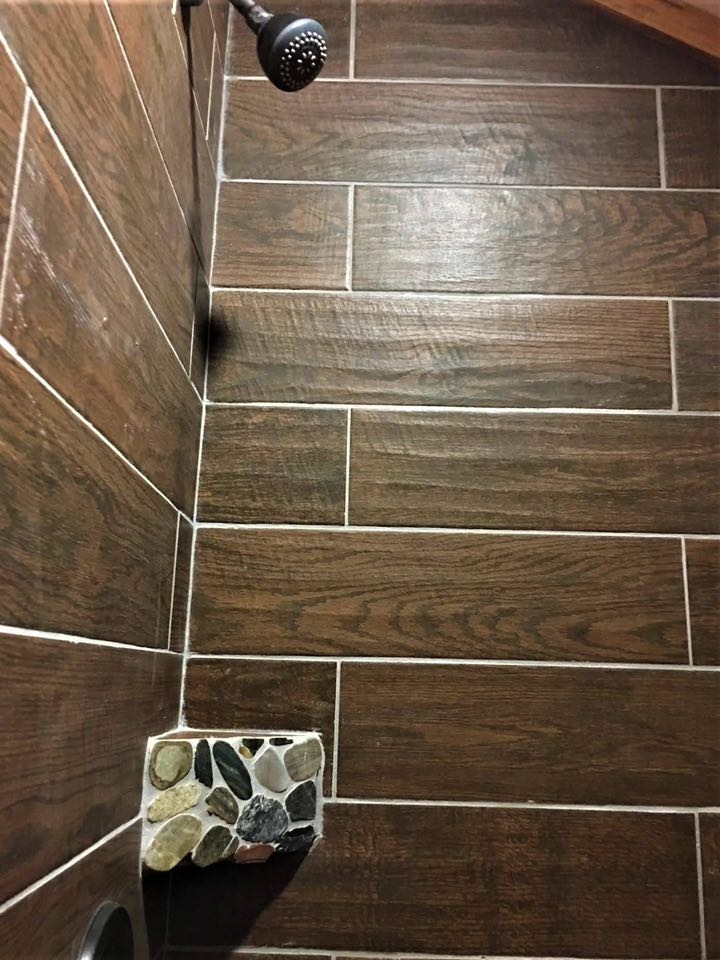 Shower wall with horizontal brown wood-like tiles, a black shower head, and a small shelf featuring multicolored stones. Earthy and minimalist design.
