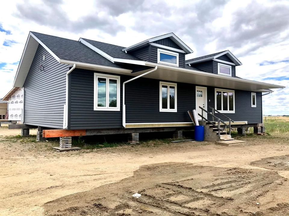 Large, dark gray house elevated on cinder blocks in a rural setting. White windows and trim, with front steps and cloudy sky above.