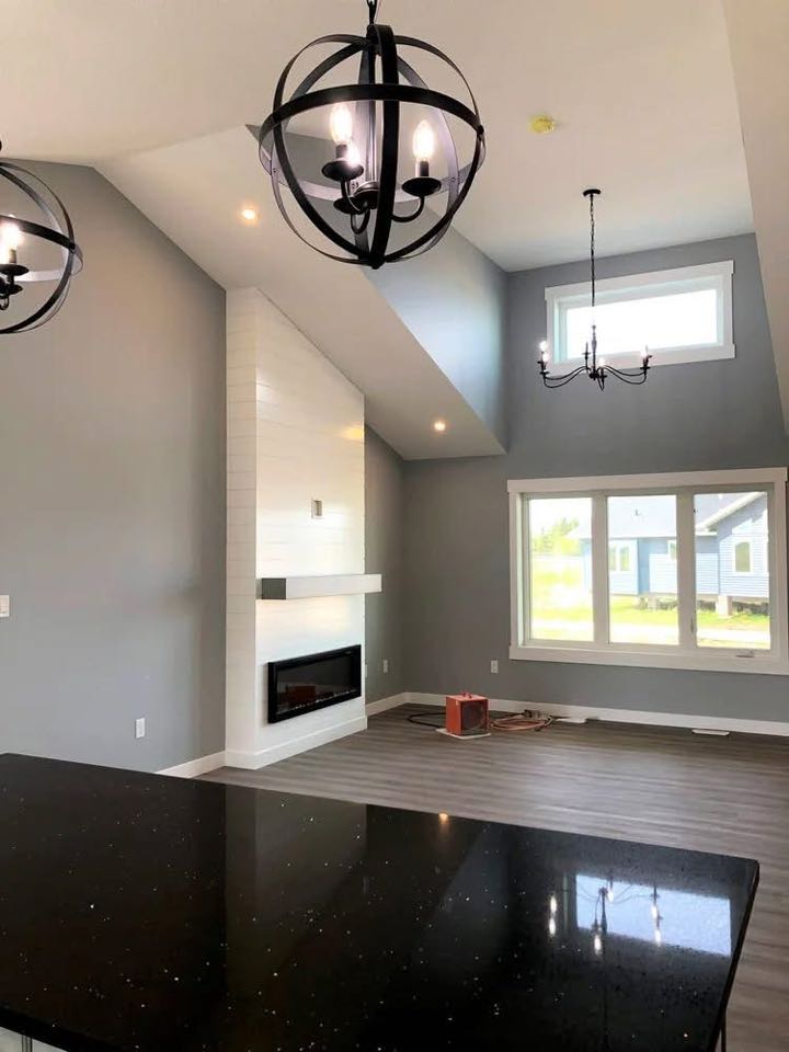 Modern living room with high ceilings, gray walls, and dark wood flooring. A sleek fireplace is adjacent to large windows. Stylish chandeliers hang above.