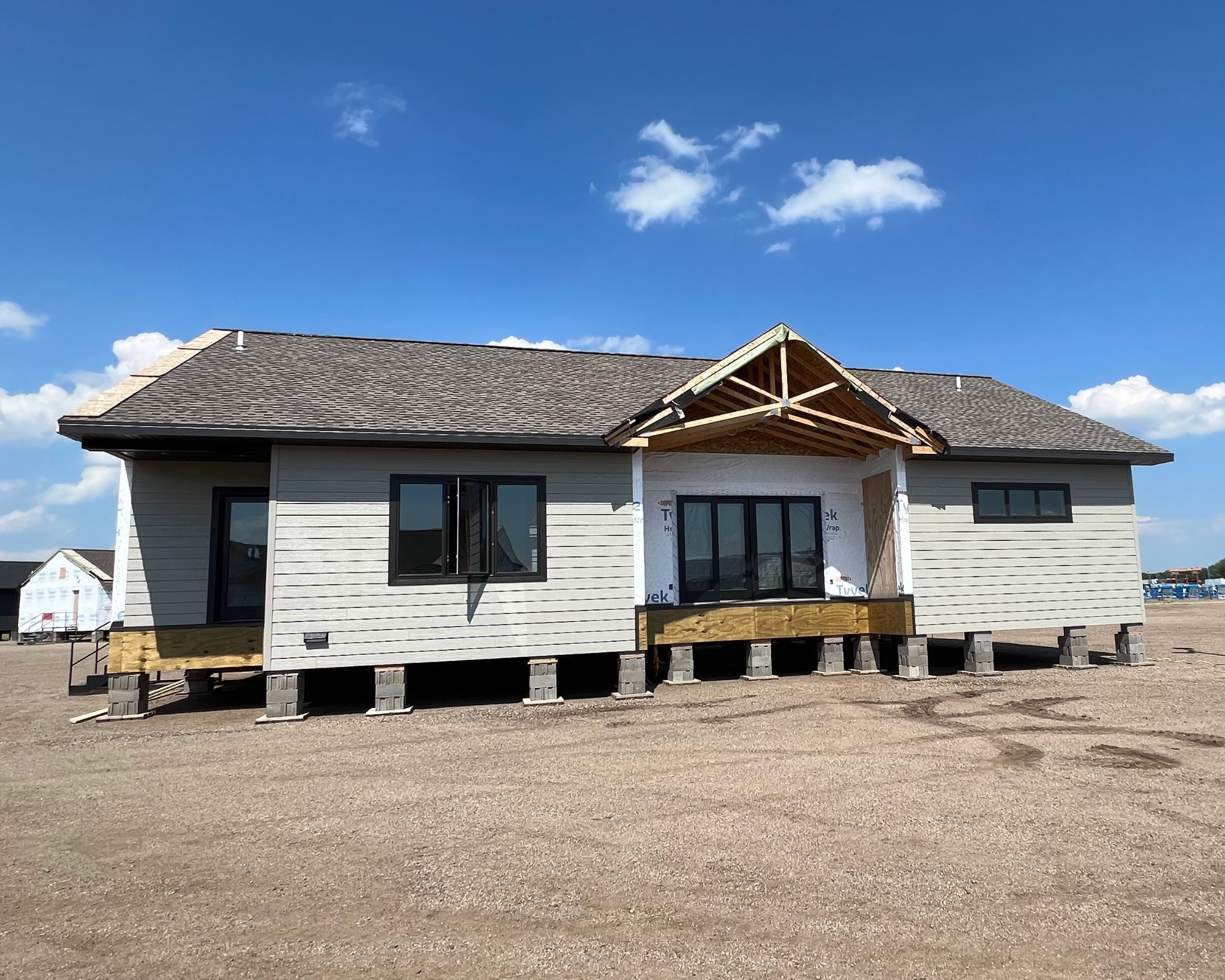 A single-story house under construction sits on a dirt lot against a clear blue sky. The structure is raised, with an incomplete porch roof visible.