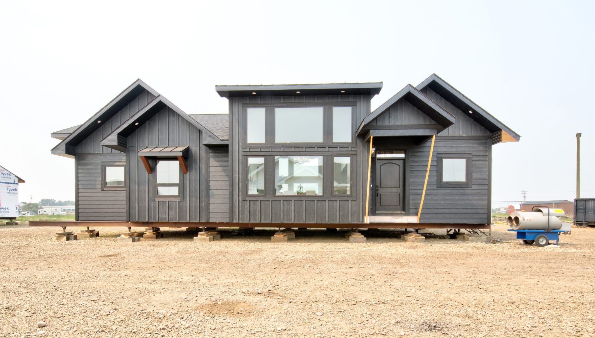 Single-story, dark gray modular home on raised blocks in an open area. Features large windows, dual gabled roofs, and a central front door, conveying modern simplicity.