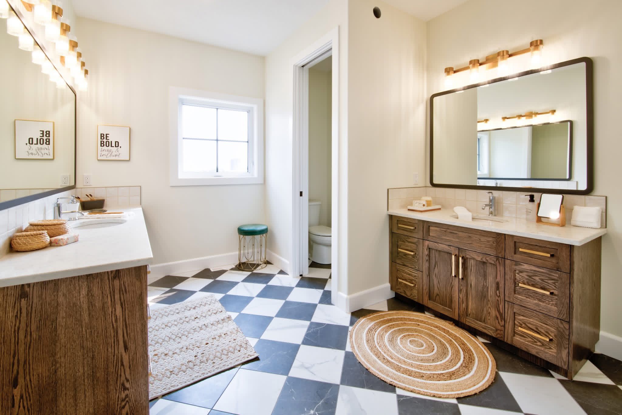 Spacious bathroom with dual wooden vanities, large mirrors, and warm lighting. Black and white checkered floor, round rug, and "Be Bold" wall art add modern flair.