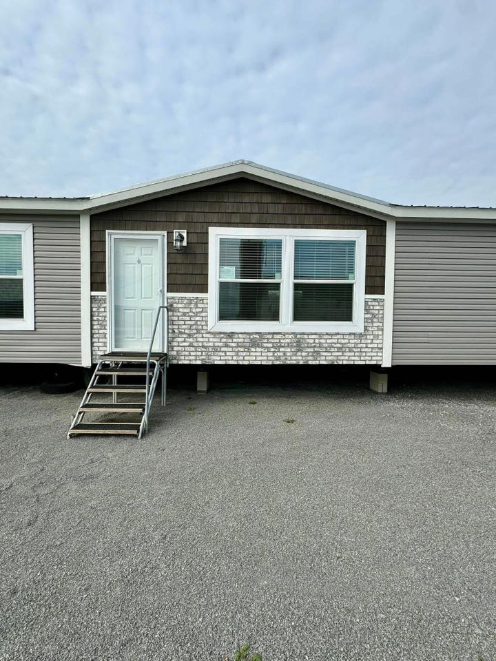 A mobile home with gray siding, a white front door, and large windows. Stairs lead to the entrance on a gravel surface, conveying a simple, functional design.