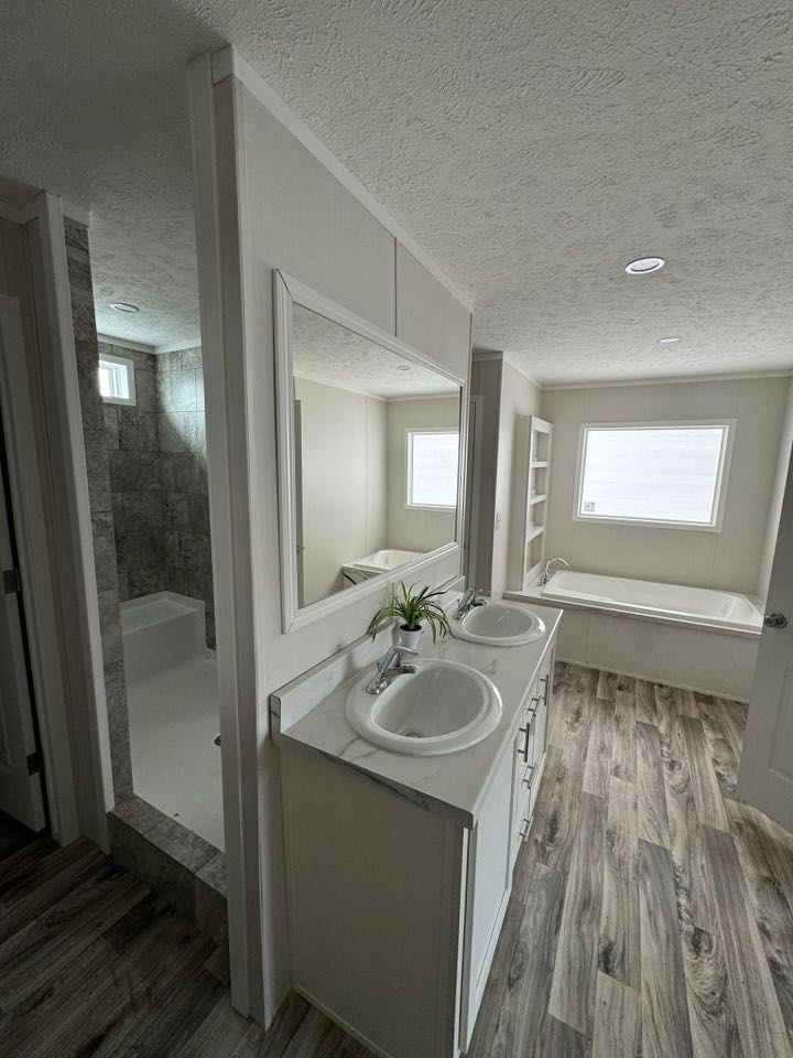 Modern bathroom featuring dual sinks with a large mirror, light wood flooring, and a soaking tub by the window. Neutral tones create a serene ambiance.