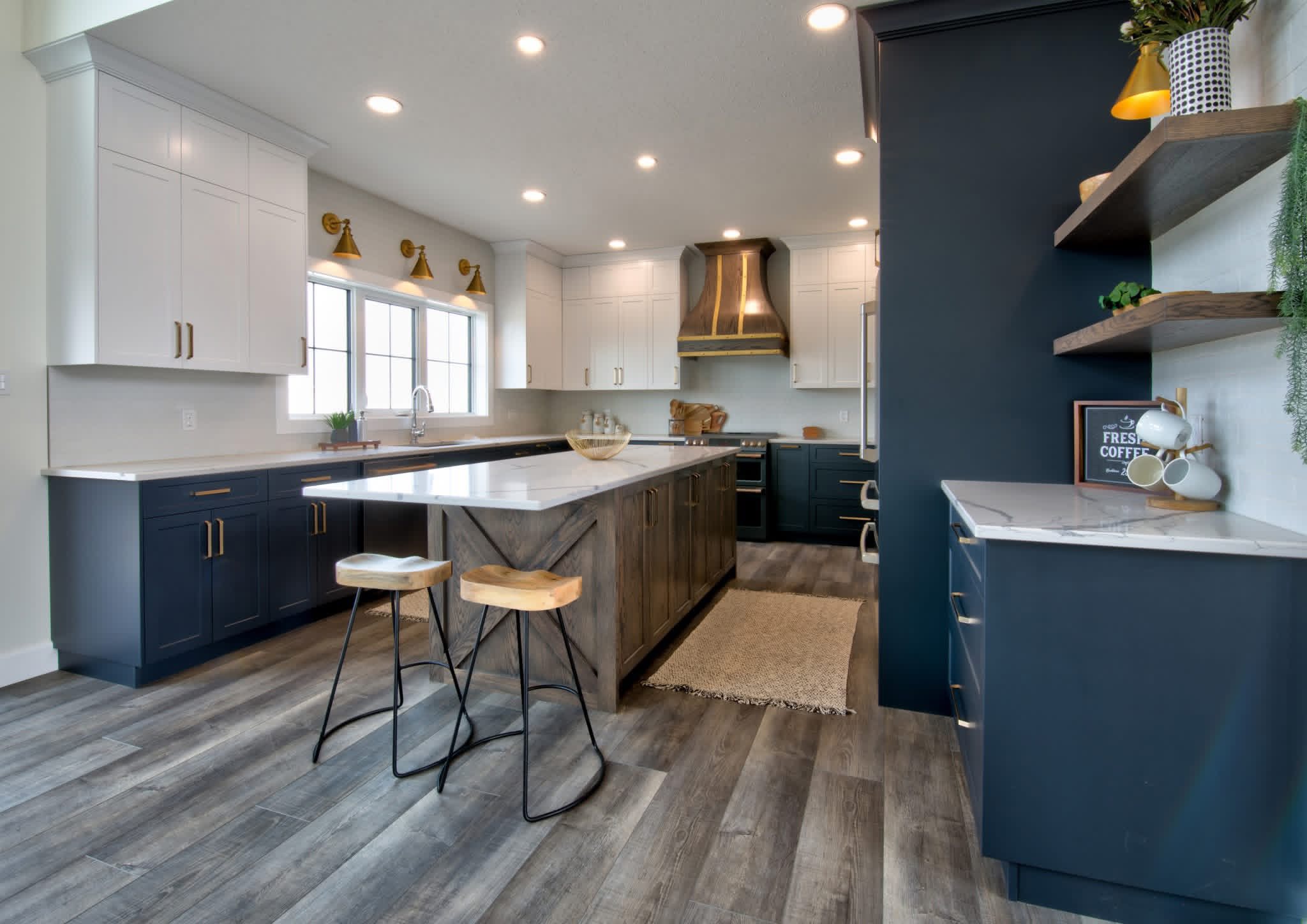 Modern kitchen with navy cabinets, white countertops, and brass fixtures. An island with wooden stools is centered on vinyl flooring under soft lighting.