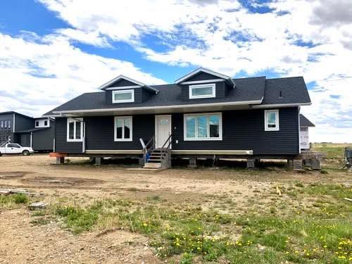 A modern, dark gray ranch-style house on a cloudy day, elevated on stilts with steps leading to the door, surrounded by open grassland.