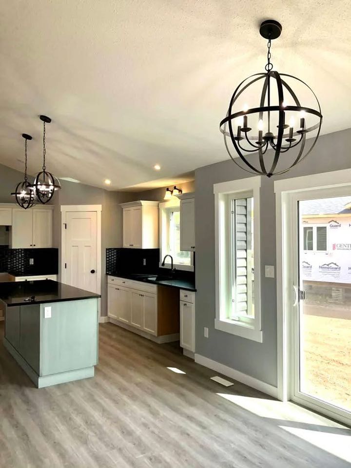 Modern kitchen with light grey walls and wood flooring. Features two black metal chandeliers, white cabinets, black countertops, and a large window. Bright and airy ambiance.