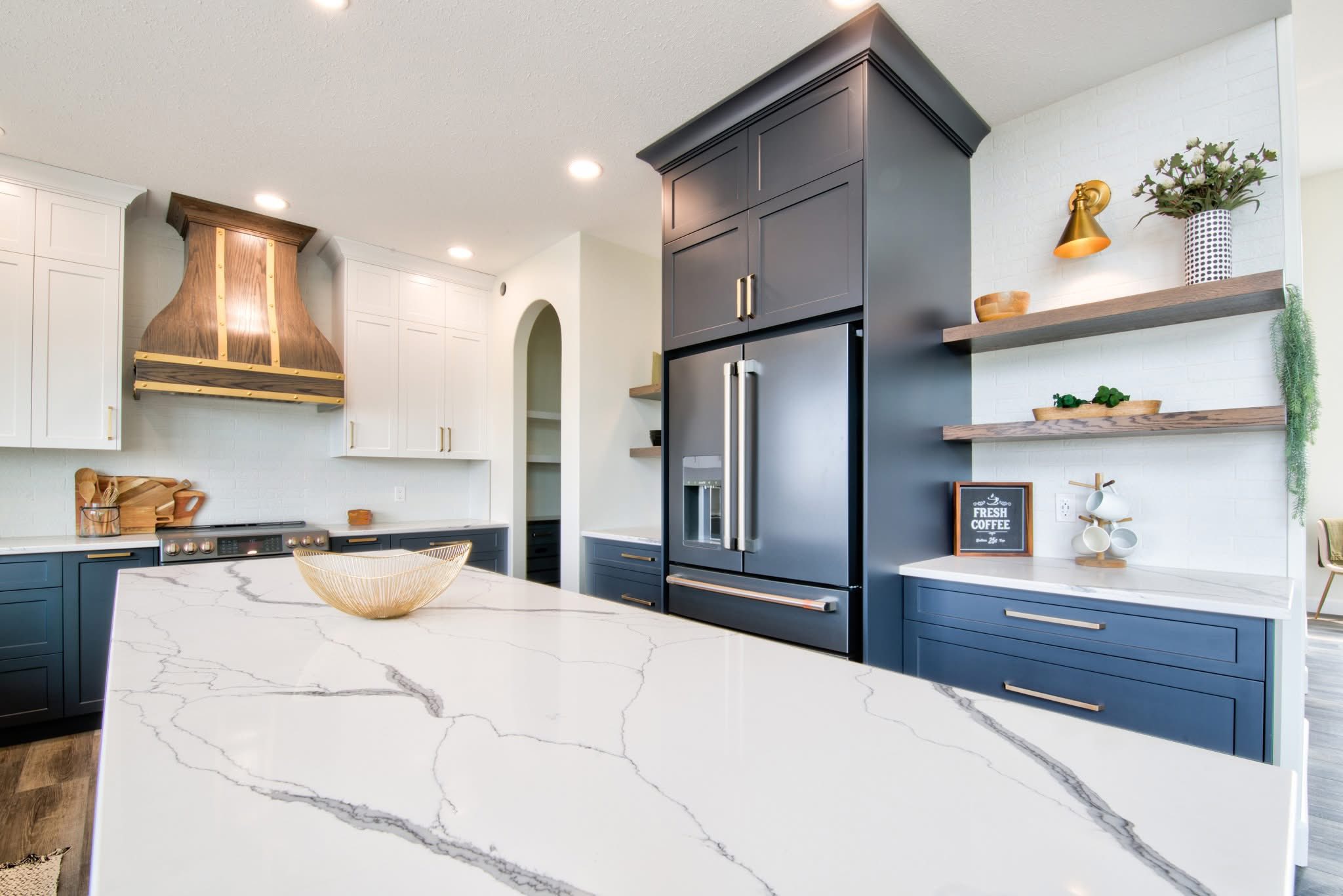 Modern kitchen with navy and white cabinets, marble island, and wooden shelves. Copper accents, stylish decor, and sleek appliances create a chic, inviting atmosphere.