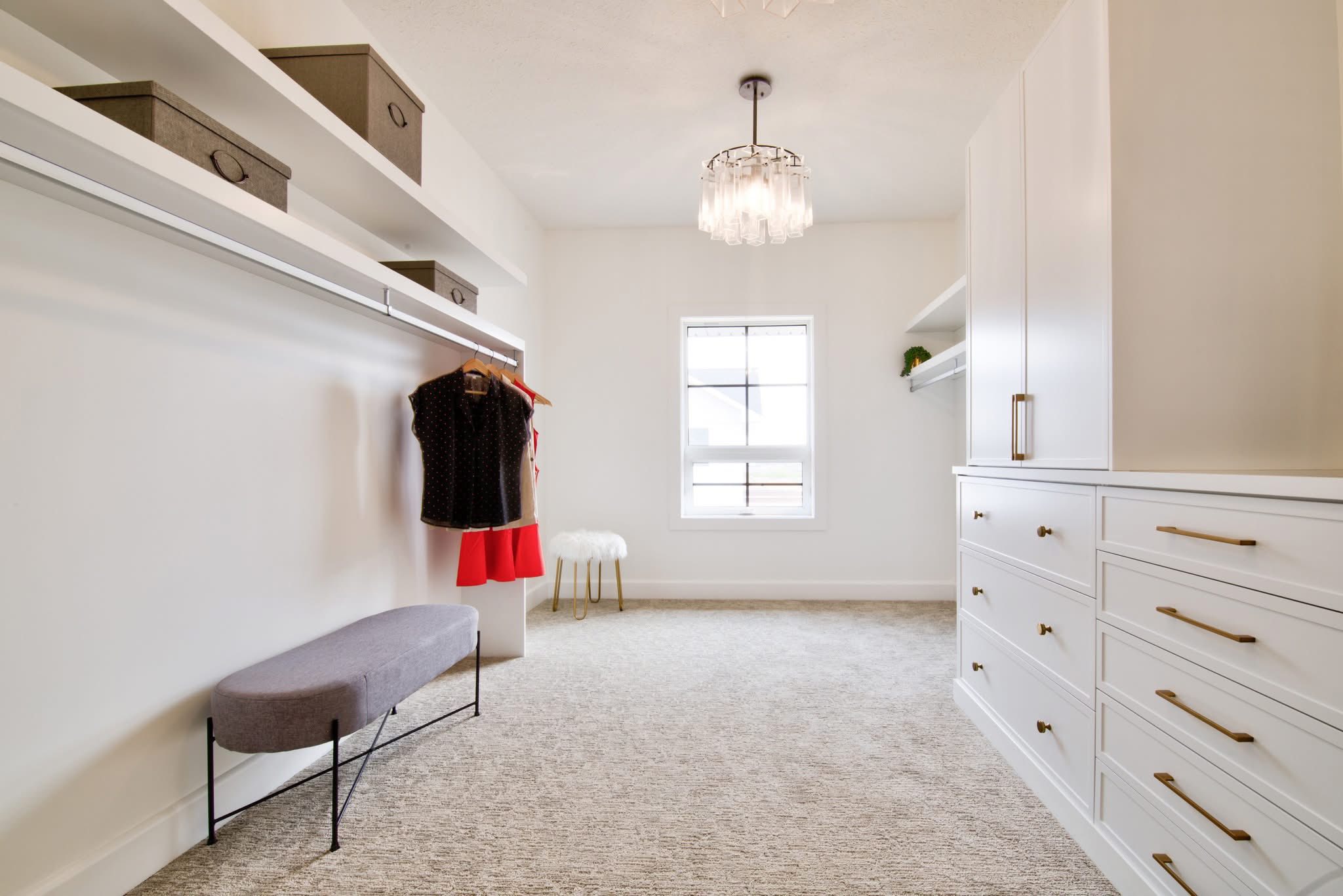 A spacious, elegant walk-in closet with white cabinets and drawers, a hanging rod with clothes, a gray bench, and a window allowing natural light.