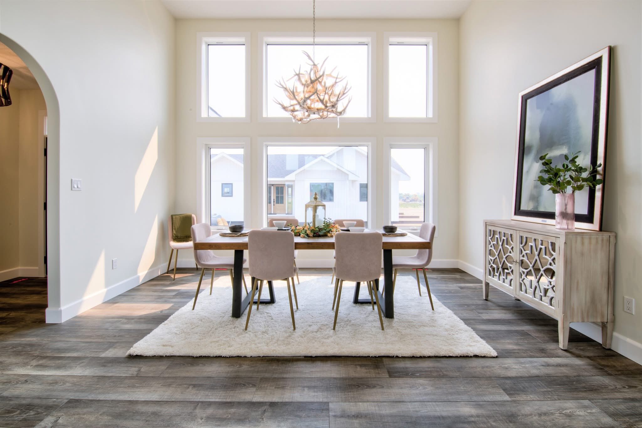 Bright dining room with large windows, a wooden table, and beige chairs on a white rug. Features a decorative sideboard, plant, and a chandelier.