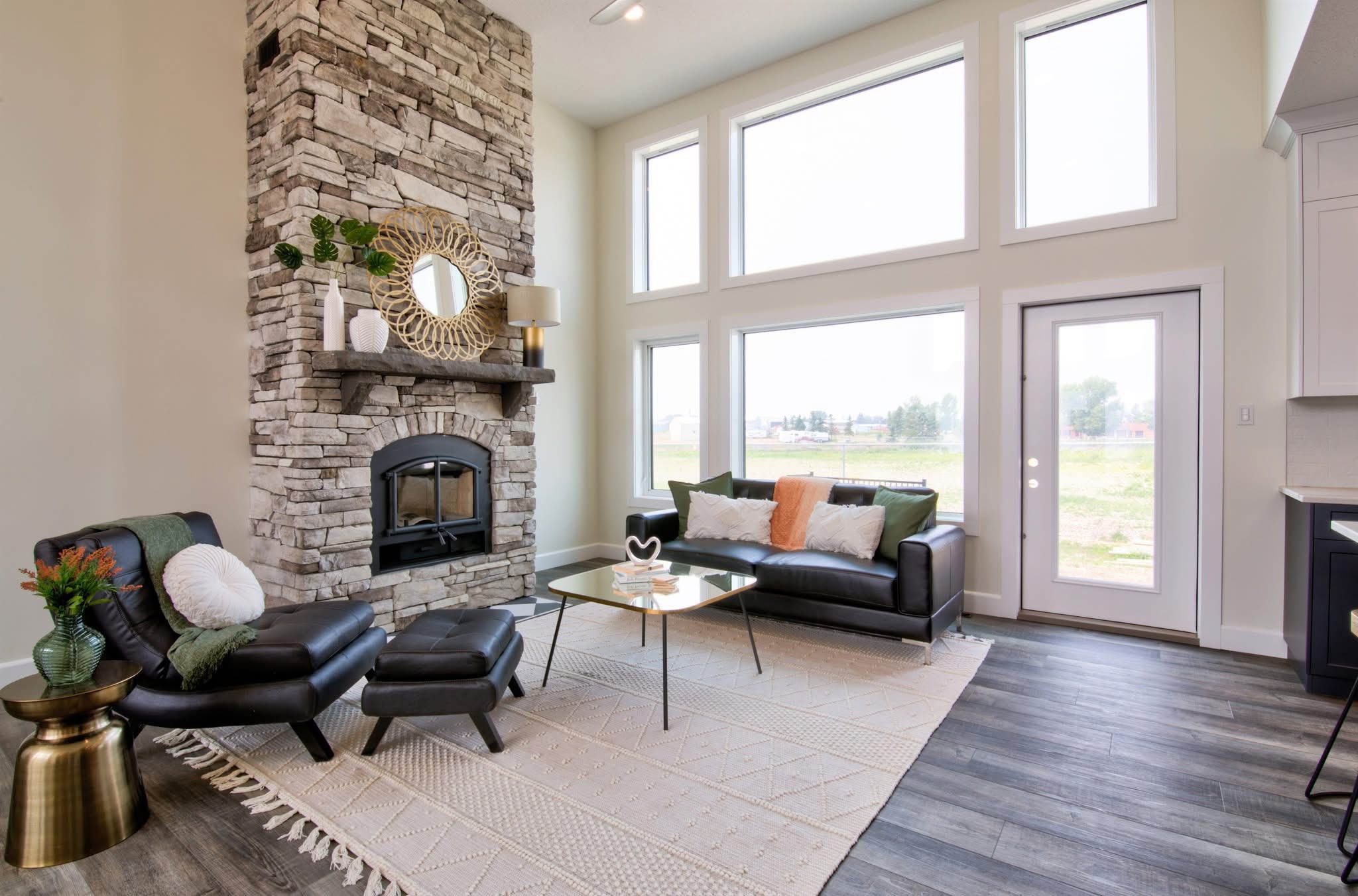 Modern living room with large windows, a stone fireplace, and sleek black furniture. Neutral tones create a cozy, inviting atmosphere.