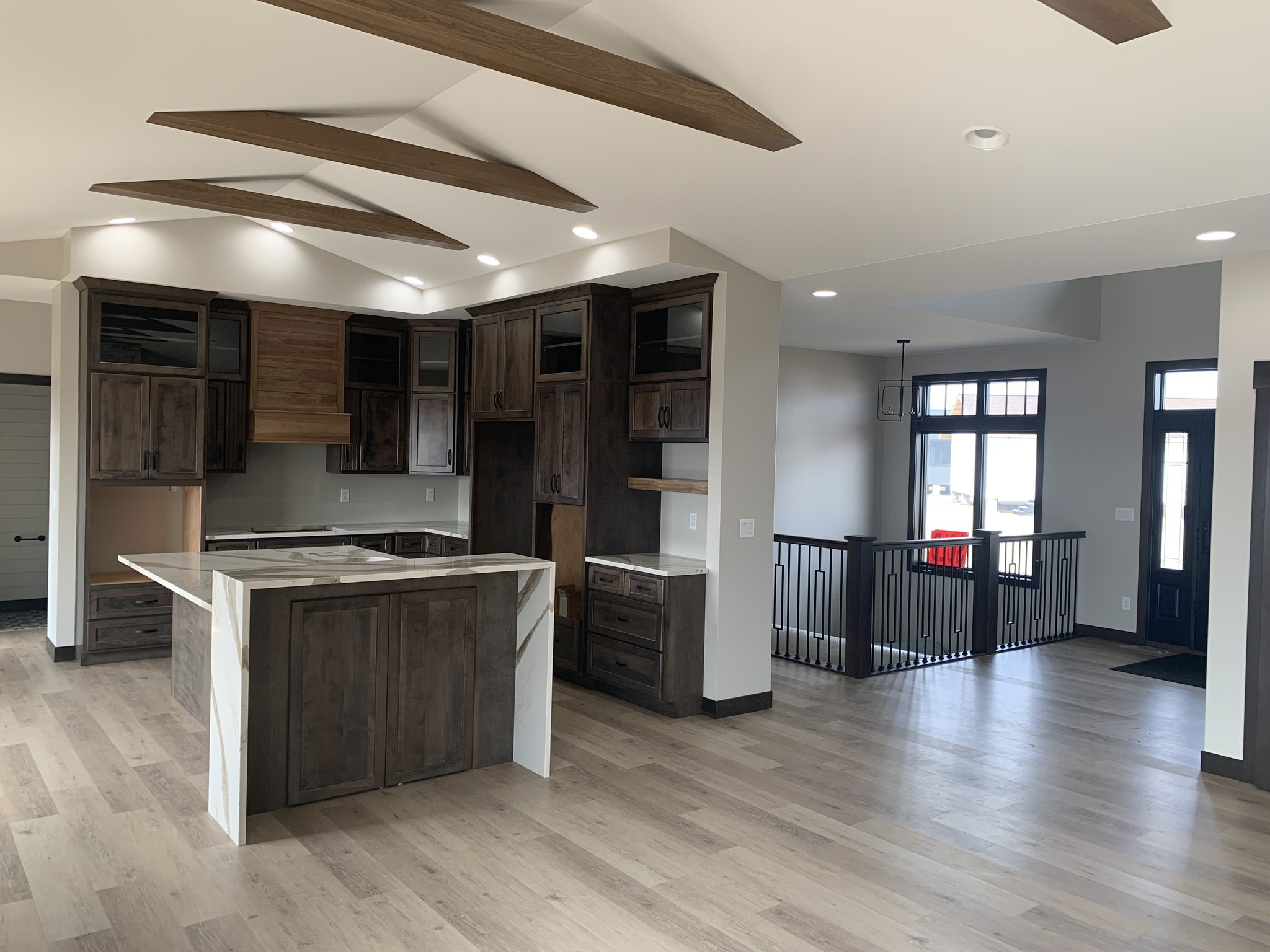Spacious modern kitchen with dark wood cabinets and an island, light wood flooring, vaulted ceiling with beams. Open layout leads to an entryway.