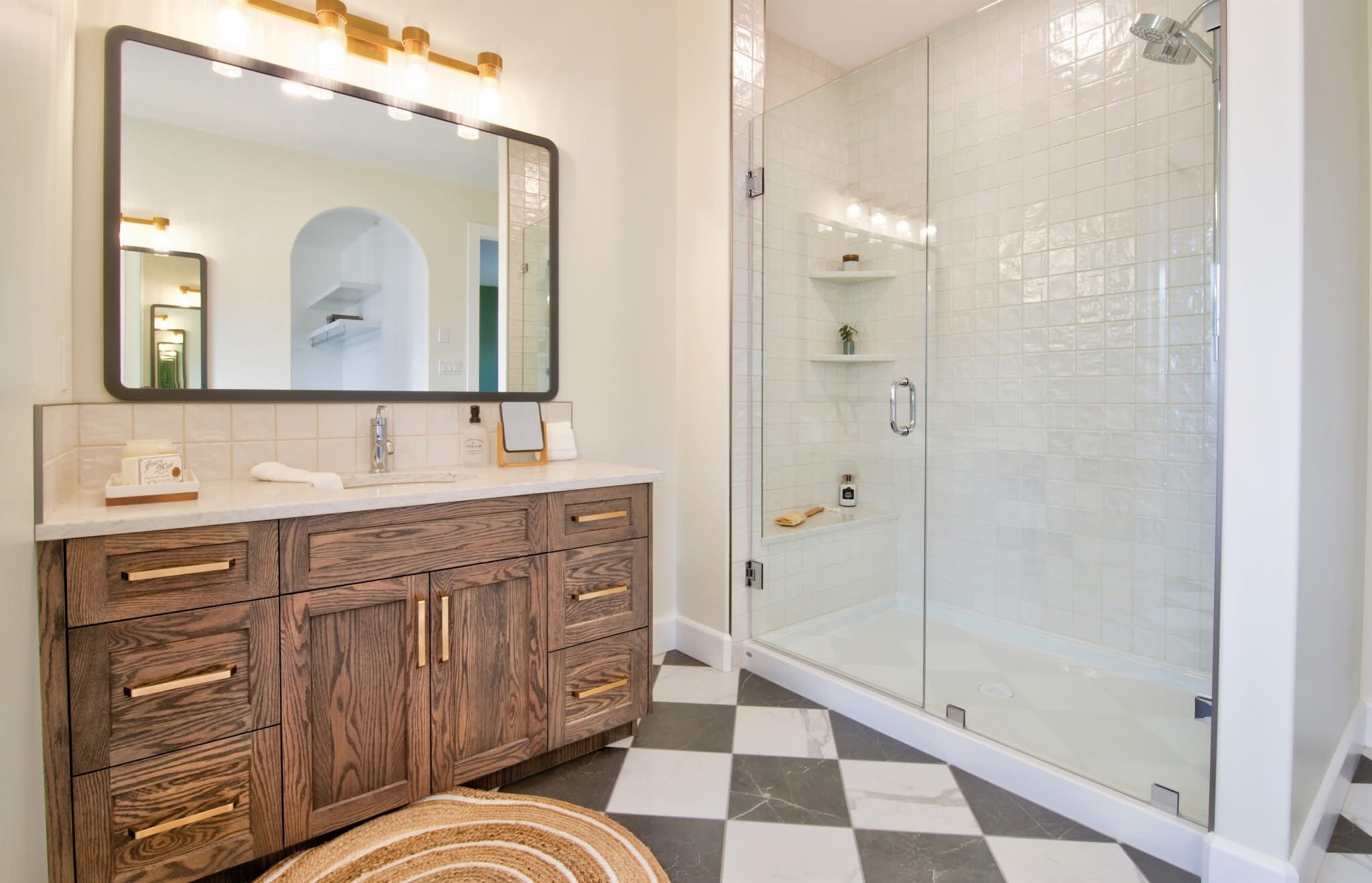 Modern bathroom with a large glass shower, wooden vanity, and black-and-white checkered floor. Warm lighting creates a cozy atmosphere.