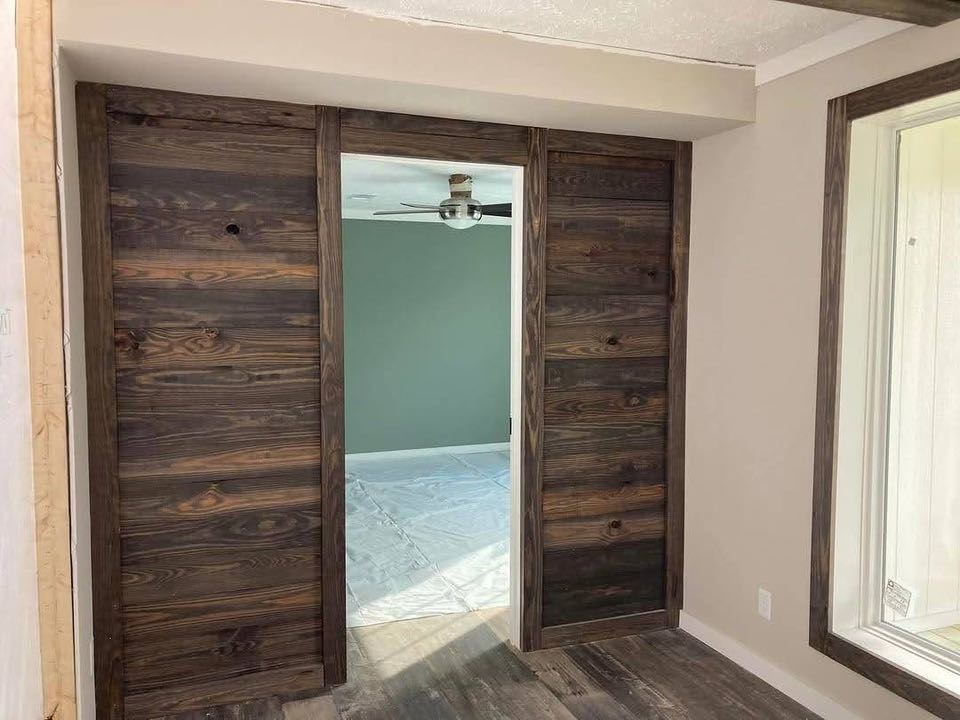 Wooden sliding barn doors open to reveal a room with sage green walls and a ceiling fan. The scene has a cozy, rustic feel.