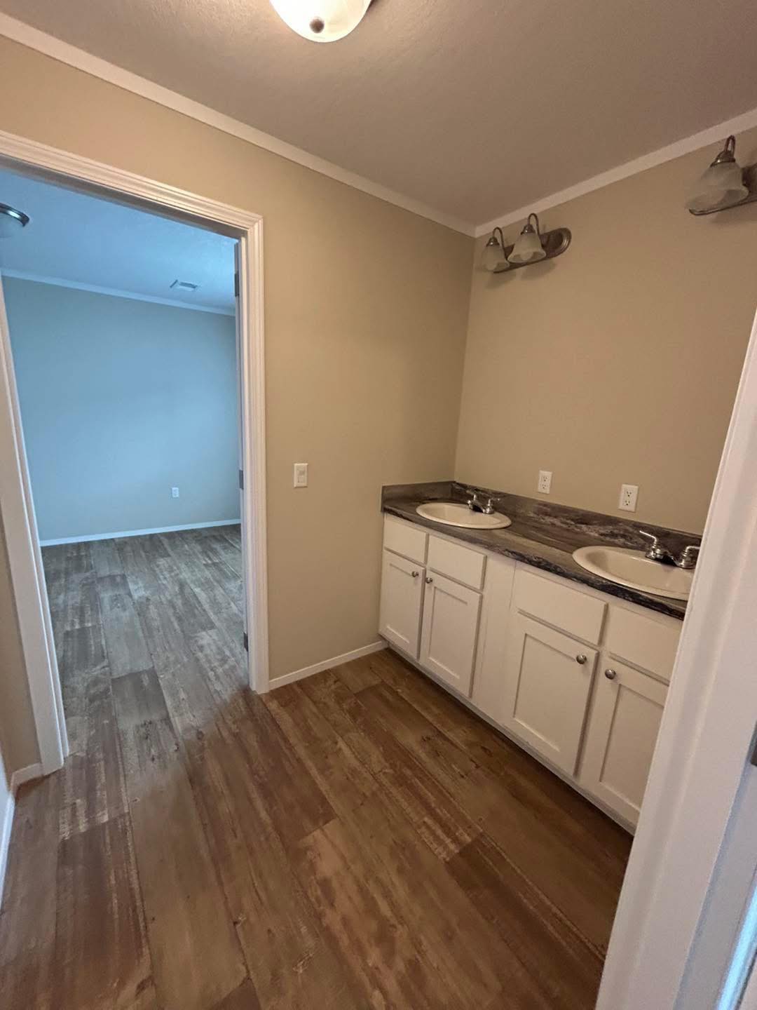 Bathroom with dual sinks on a wooden vanity, marble countertop, under warm lighting. Open doorway leads to a room with wooden flooring. Neutral tones.