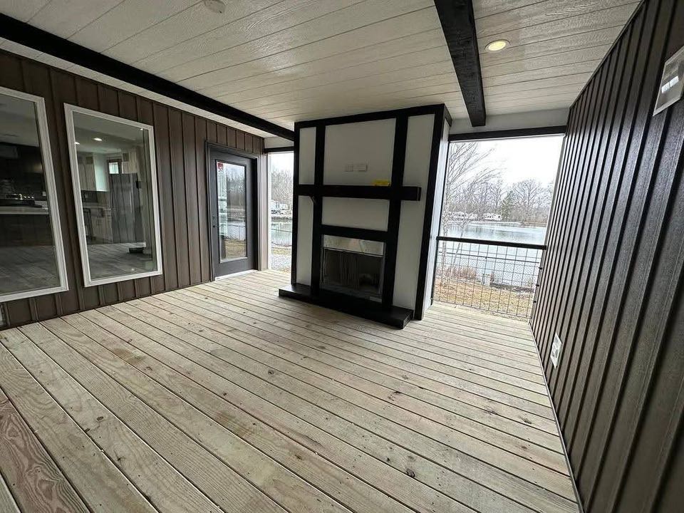 Enclosed porch with wood panels, large windows, and a sliding glass door. Features a modern fireplace and views of trees and a body of water outside.