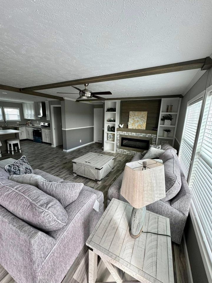 Modern living room with two gray sofas, a wooden coffee table, and a table lamp. A fireplace and shelves adorn the wall. Open kitchen view. Bright and cozy ambiance.