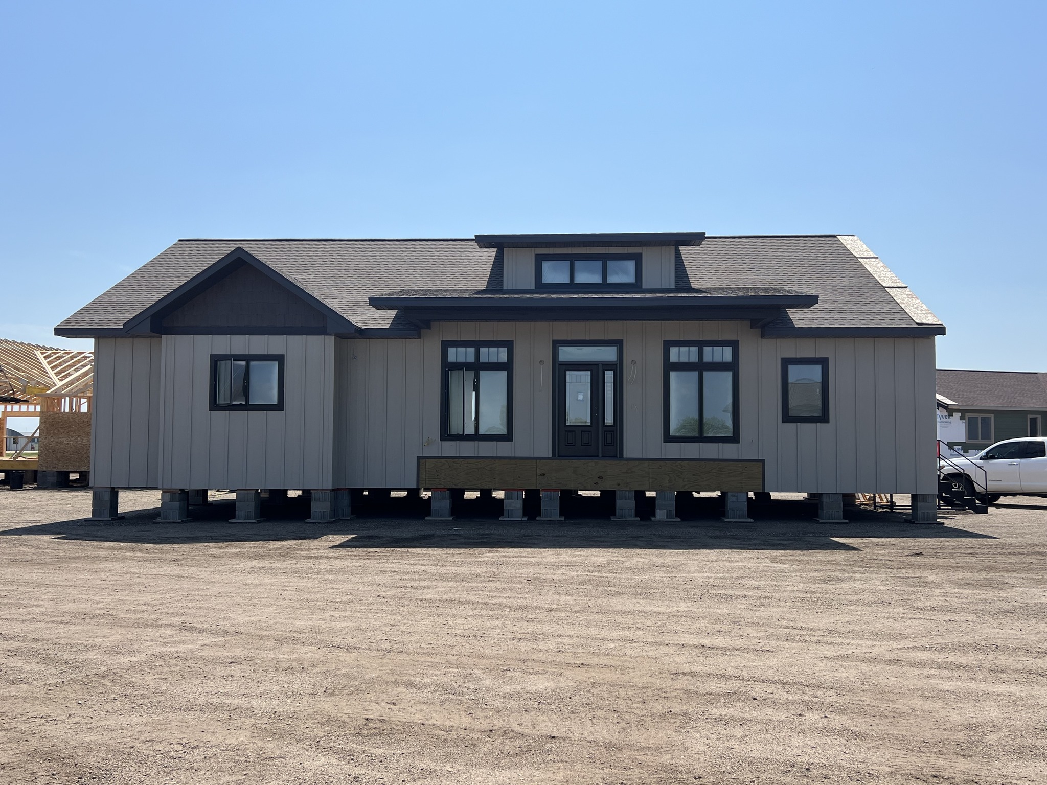 A beige prefabricated house with dark trim sits elevated on a dirt lot under a clear blue sky. The structure has multiple windows and a centered door.