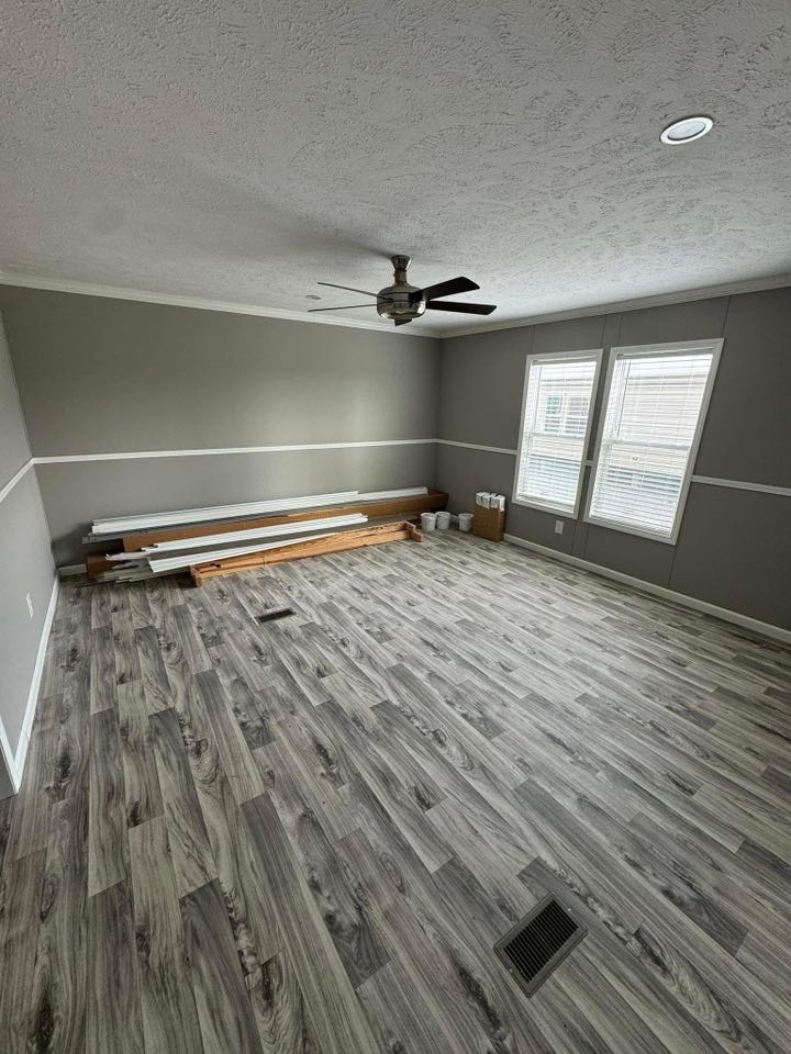 Empty room with gray laminate flooring and gray walls, featuring two large windows on the right. Construction materials and tools are neatly stacked against one wall, and a ceiling fan is mounted above.