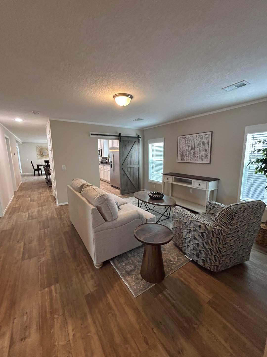 A cozy living room with a beige sofa, patterned armchair, and round wooden table on a rug. Barn door reveals kitchen, conveying a warm, inviting atmosphere.