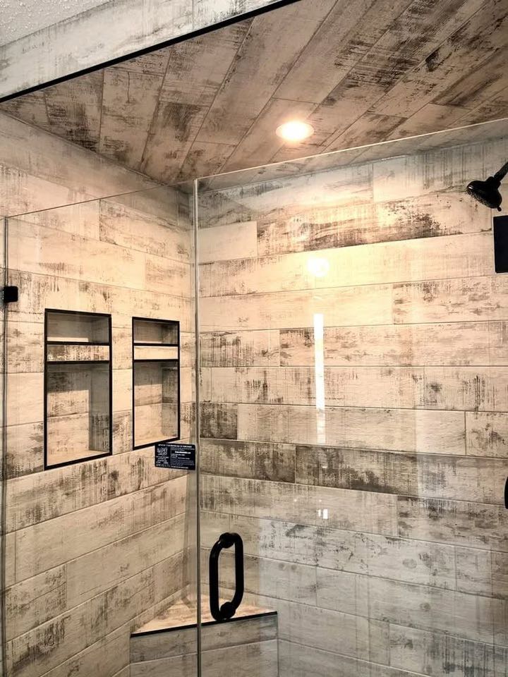 Modern shower with rustic wood-look tiles in shades of beige and gray. Glass door, black metal handle, and small recessed shelves create a sleek, cozy atmosphere.