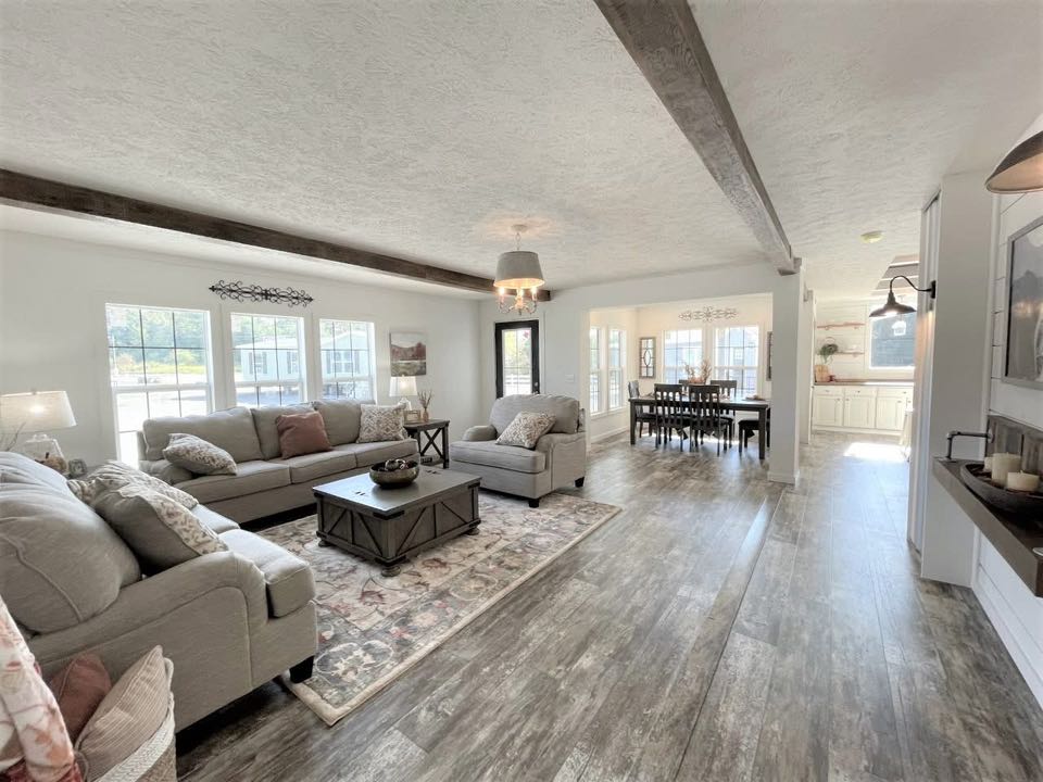 Spacious open-concept living room with light gray sofas, rustic coffee table on patterned rug, and dining area. Wooden beams and large windows add warmth.