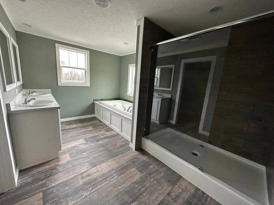 A modern bathroom with wood flooring, a double sink vanity, large glass shower, and a soaking tub by two windows. The walls are painted green.