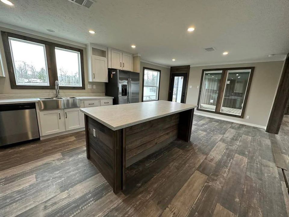 Modern kitchen with a large island, dark wood flooring, and stainless steel appliances. Bright light through large windows creates an inviting feel.
