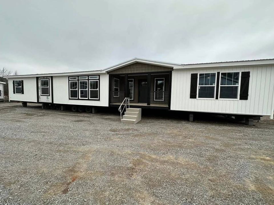 A large, white mobile home sits on a gravel lot under an overcast sky. It features multiple windows, dark trim, and a small front porch with stairs.