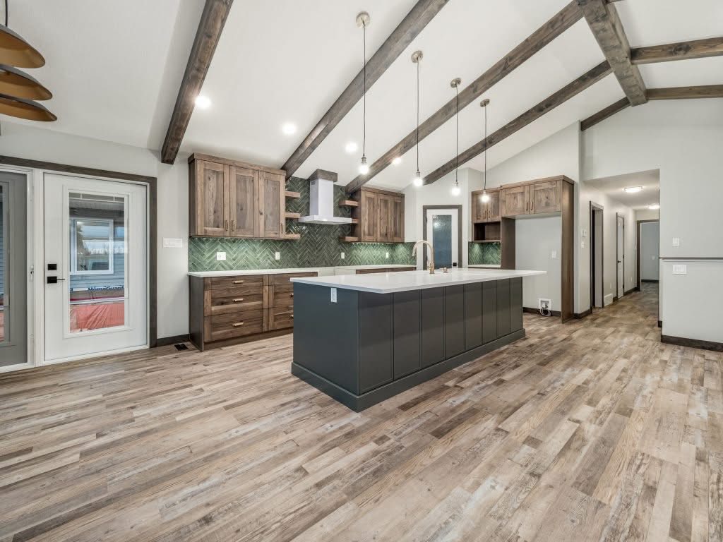 Modern kitchen with vaulted ceiling and exposed wooden beams. Features wooden cabinets, green backsplash, large island, and pendant lights, creating a warm, rustic ambiance.
