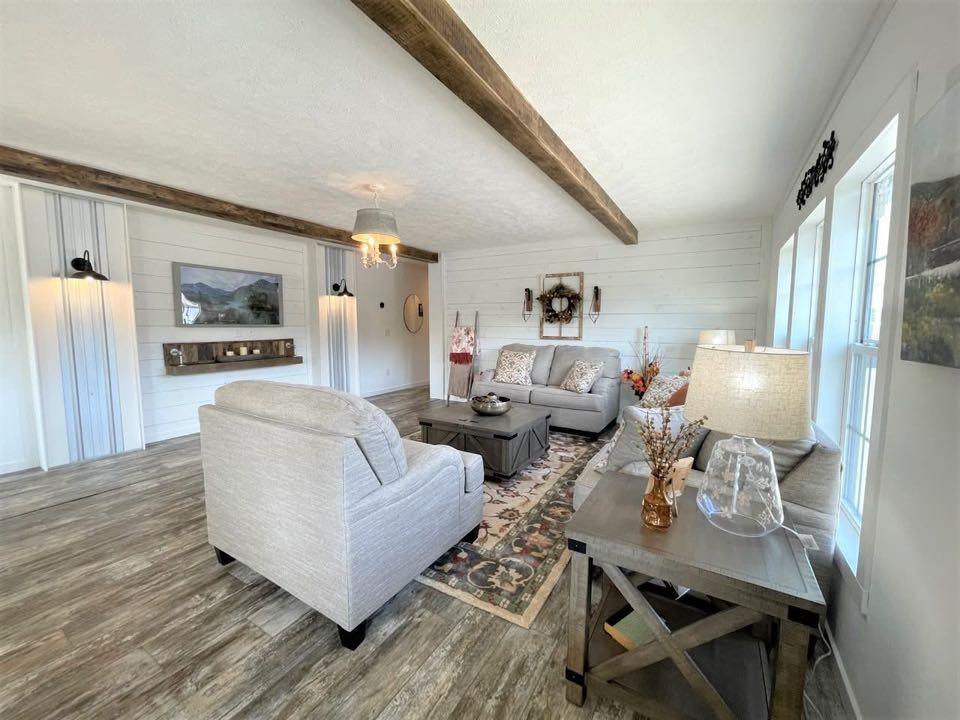 Cozy farmhouse-style living room with gray sofas, rustic wood beams, and a floral rug. Soft lighting from lamps and a chandelier creates a warm ambiance.