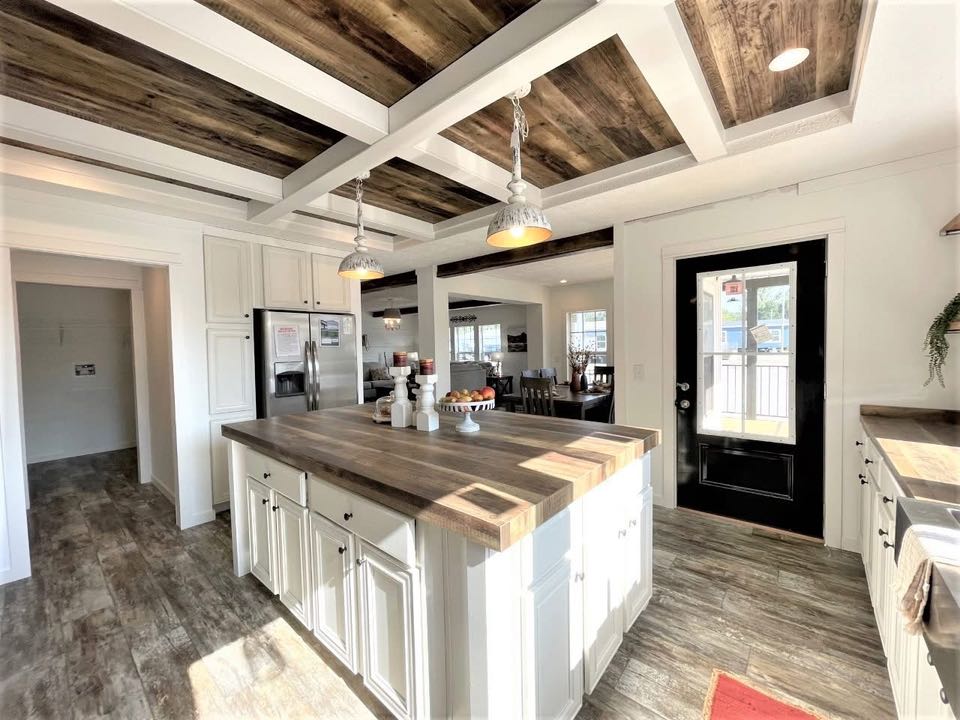 Bright kitchen featuring a large wooden island, pendant lights, white cabinets, and a dark glass door. Rustic wood floors and ceiling add warmth.