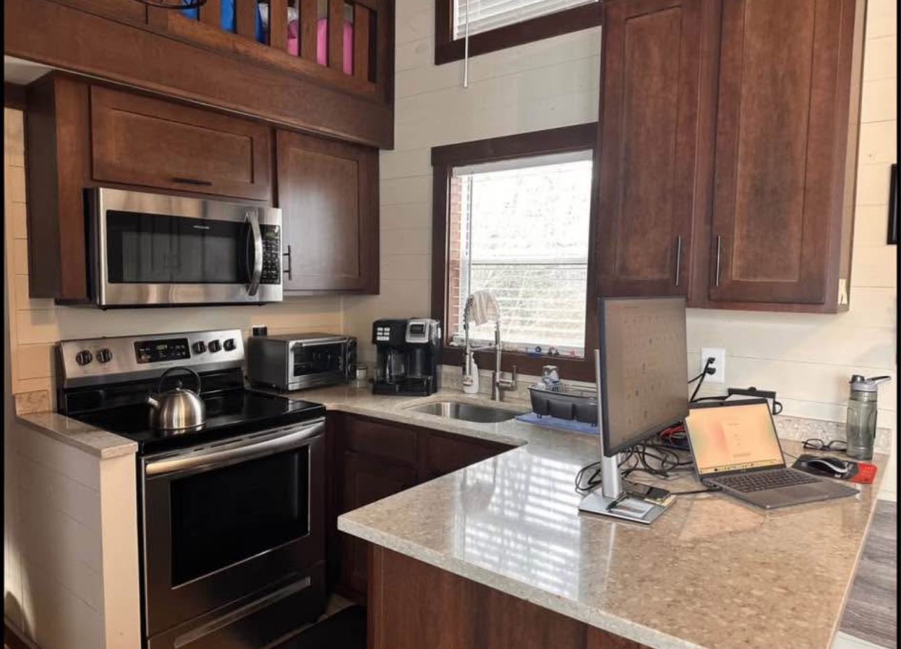 Compact kitchen with dark wooden cabinets, stainless steel appliances, a laptop on the counter, and streamed sunlight from a window, creating a cozy atmosphere.