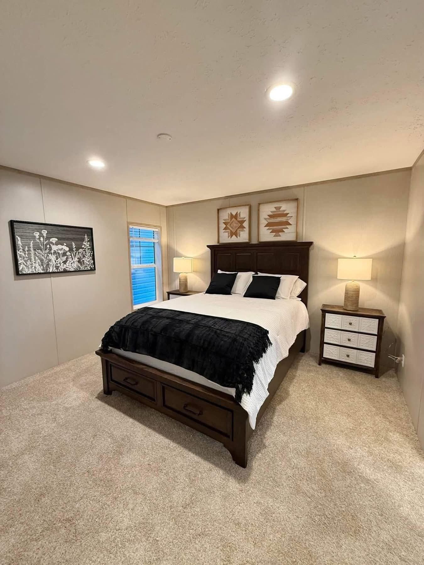 Cozy bedroom with a dark wood bed, white and black bedding, and two bedside lamps. Soft beige carpet, floral artwork, and warm lighting create a calm ambiance.