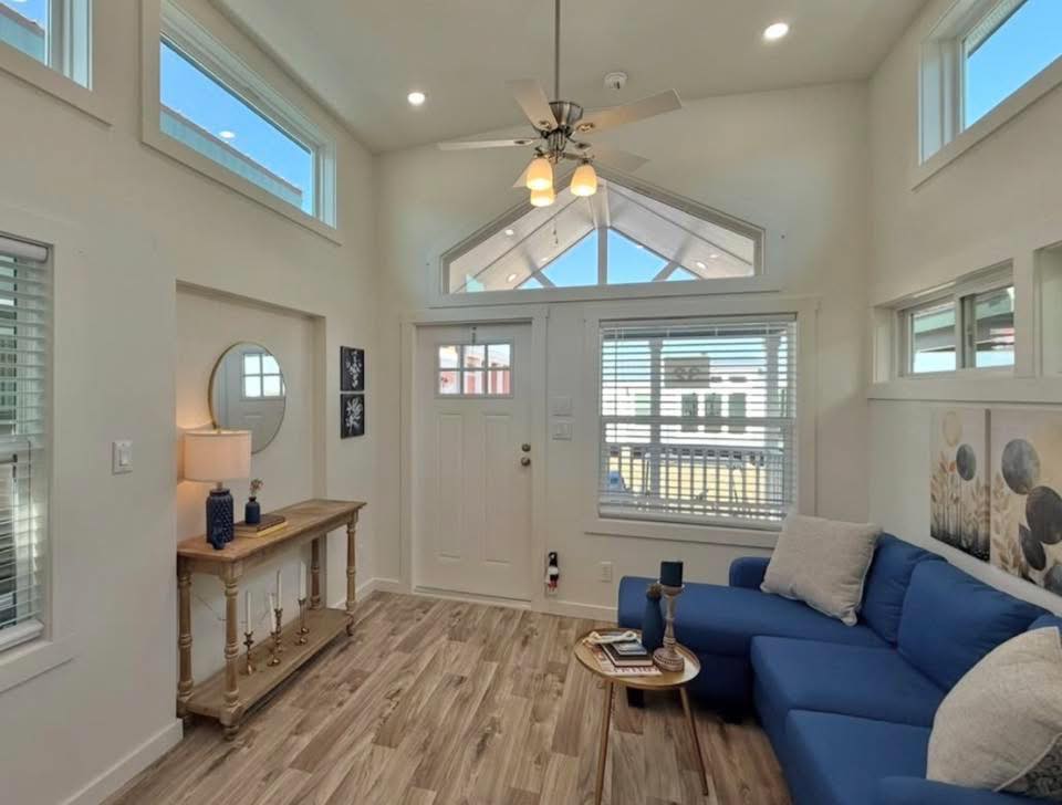 Bright, airy living room features wood floors, a blue couch, coffee table, and decor accents. Large windows and white walls enhance natural light.