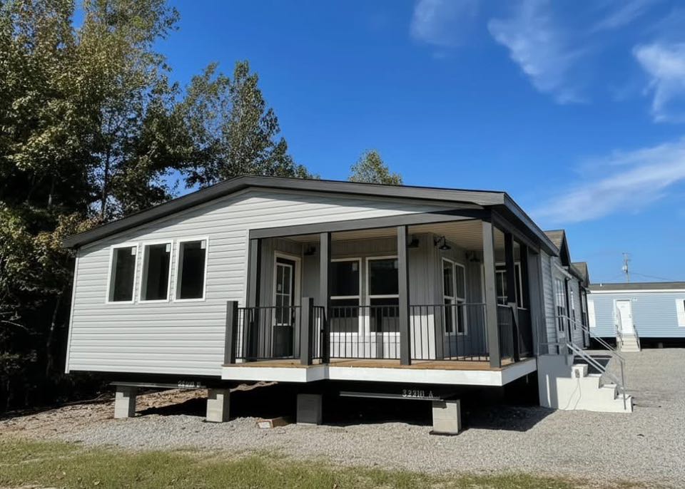 Single-story modular home with white siding, a covered front porch, and black railings. Located on a gravel lot, surrounded by trees under a blue sky.