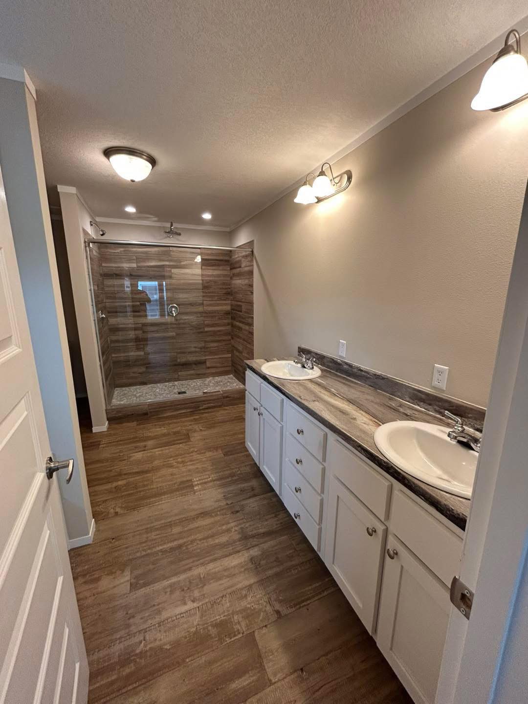 Modern bathroom with wooden flooring and beige walls. Features a double sink with marble countertop on the right and a spacious glass-enclosed shower at the end. Bright ceiling lights create a clean, elegant ambiance.