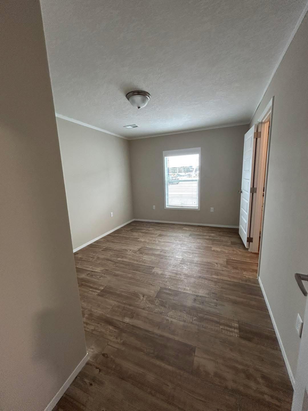A small, empty room with light beige walls and wooden flooring. A single window lets in natural light, and a closed white door is on the right.